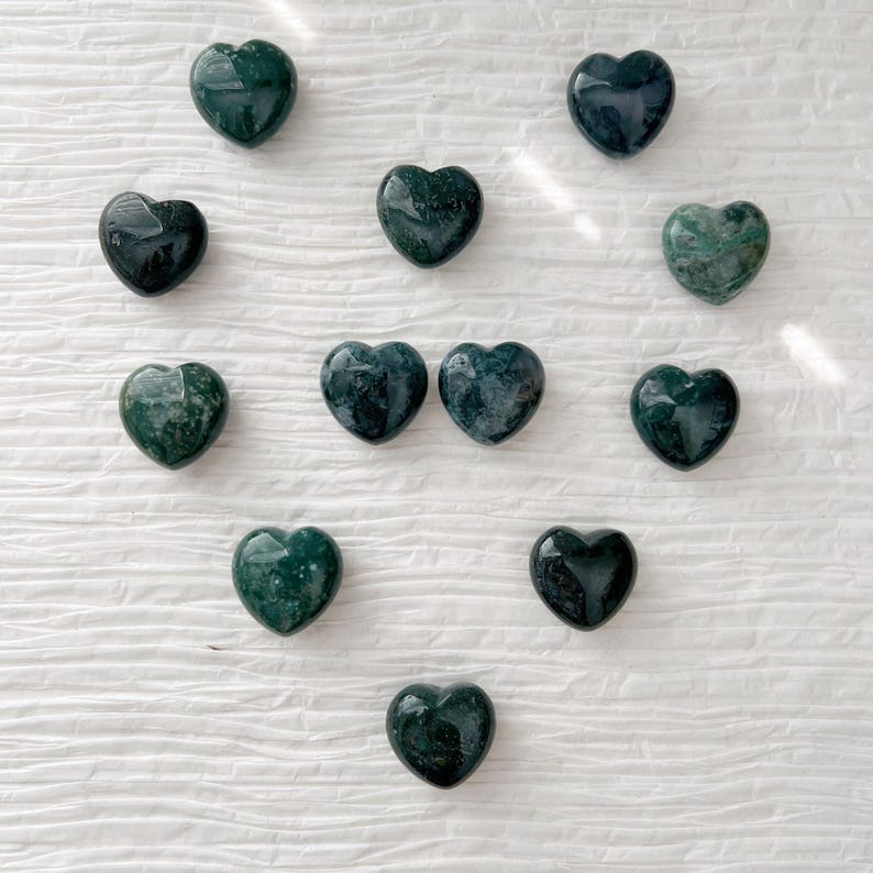 May include: A collection of heart-shaped stones in varying shades of green and dark teal. The stones have a polished, smooth surface and are arranged in a scattered pattern. The stones appear to be made of a natural material.