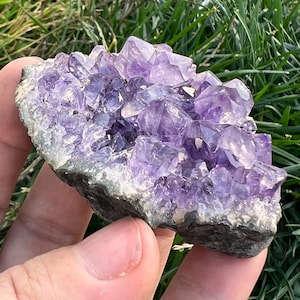 May include: A cluster of amethyst crystals in a natural formation. The crystals are a deep purple color and have a smooth, polished surface. The amethyst cluster is attached to a gray, rocky base.