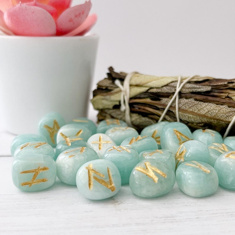 May include: A set of 20 teal blue tumbled stones with gold runes inscribed on each. The stones are scattered on a white wooden surface.