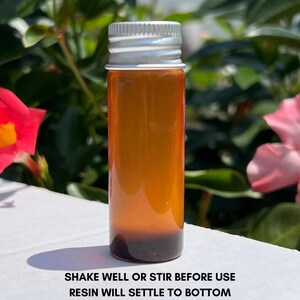 May include: A small glass bottle with a silver lid, containing a dark amber liquid. The text "SHAKE WELL OR STIR BEFORE USE RESIN WILL SETTLE TO BOTTOM" is printed in black at the bottom. The background shows green leaves and pink flowers.
