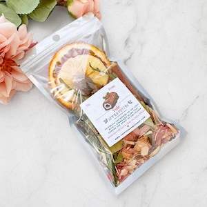 May include: A clear plastic bag filled with dried botanicals, including orange slices, cinnamon sticks, and herbs. A small tag reads "Yule Simmer Pot Mix" with text about celebrating the season. The bag is sealed and ready for use.