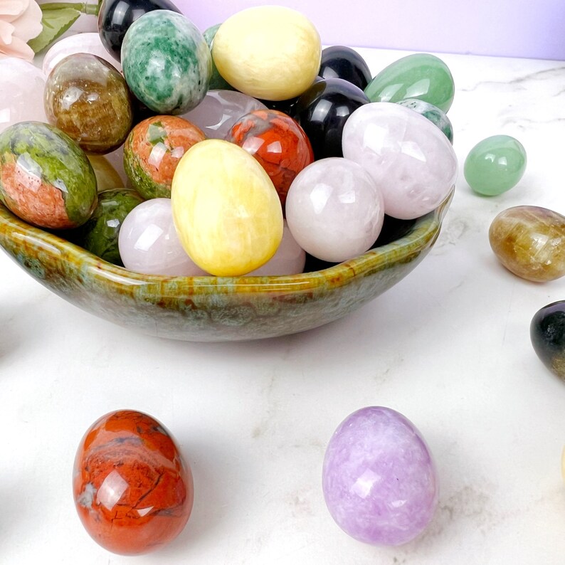 Puede incluir: Un bol lleno de huevos de piedras preciosas pulidas de varios colores, incluyendo verde, rosa, amarillo, rojo y marr&oacute;n. Los huevos est&aacute;n dispuestos en un bol con un esmalte verde y marr&oacute;n.