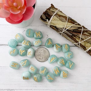 May include: A set of 20 teal green tumbled stones with gold runes painted on them. The stones are arranged in a circle on a white surface.