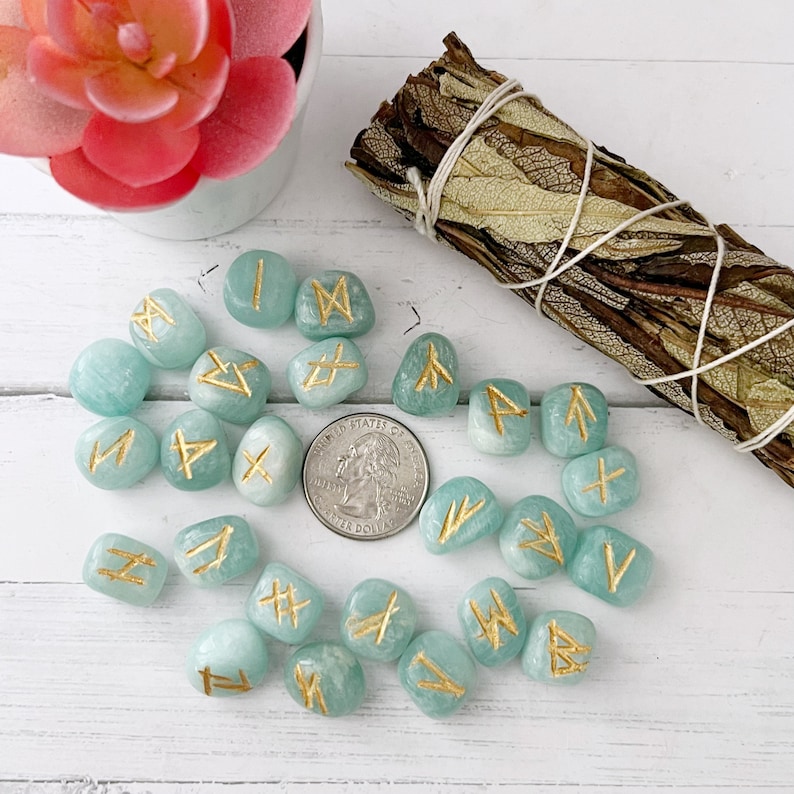 May include: A set of 20 teal green tumbled stones with gold runes painted on them. The stones are arranged in a circle on a white surface.
