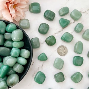 May include: A collection of tumbled green aventurine stones. The stones are smooth and polished, and they are arranged in a bowl and scattered on a white surface.