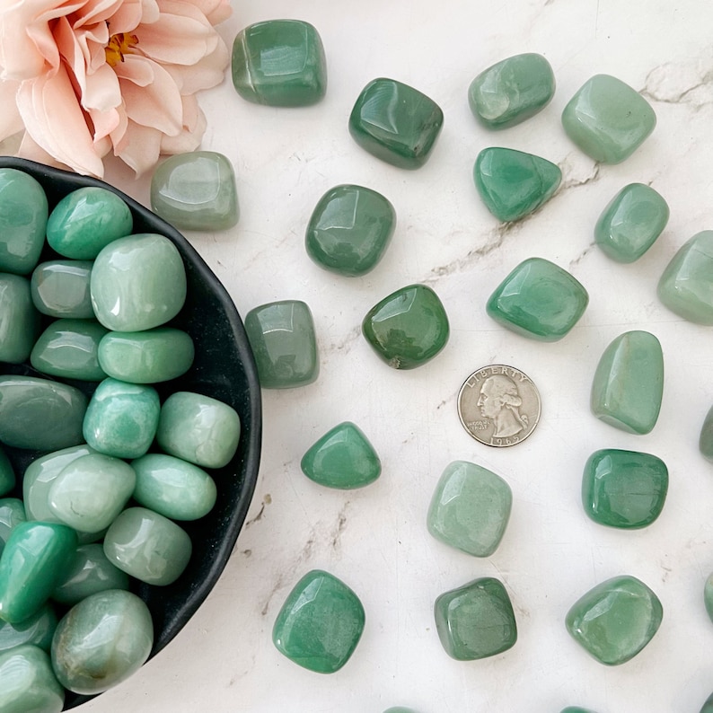May include: A collection of tumbled green aventurine stones. The stones are smooth and polished, and they are arranged in a bowl and scattered on a white surface.