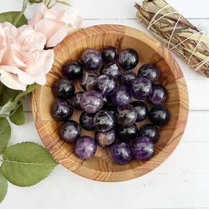 May include: A wooden bowl filled with amethyst spheres. The spheres are a deep purple color with some having a lighter, almost white, center. The bowl is sitting on a white surface.