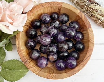 Puede incluir: Un cuenco de madera lleno de esferas de amatista. Las esferas son de color morado oscuro, algunas tienen un centro más claro, casi blanco. El cuenco está sobre una superficie blanca.