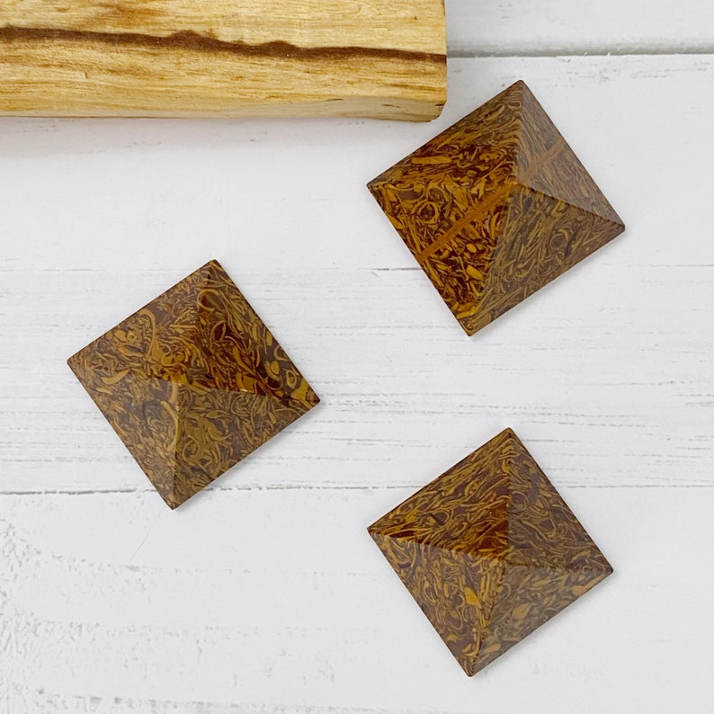 May include: Three brown and gold patterned stone pyramids with a square base and pointed top are arranged on a white wooden surface. A light brown wooden stick is in the background. The pyramids have a unique, natural pattern.