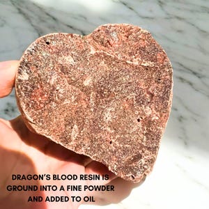 May include: A heart-shaped piece of dragon's blood resin, reddish-brown in colour, held in a hand. The resin has a textured surface with small holes. Text on the image reads: "DRAGON'S BLOOD RESIN IS GROUND INTO A FINE POWDER AND ADDED TO OIL."