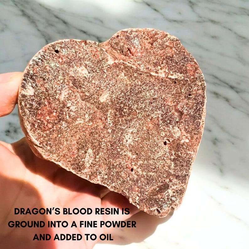 May include: A heart-shaped piece of dragon's blood resin, reddish-brown in colour, held in a hand. The resin has a textured surface with small holes. Text on the image reads: "DRAGON'S BLOOD RESIN IS GROUND INTO A FINE POWDER AND ADDED TO OIL."