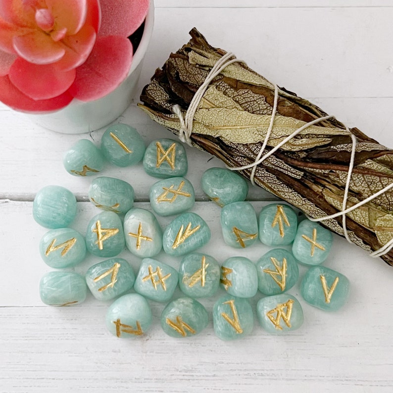 May include: A set of 20 teal green tumbled stones with gold runes painted on them. The stones are arranged on a white surface with a bundle of dried sage in the background.