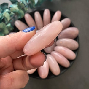 Pu&ograve; includere: Una collezione di pietre di quarzo rosa ovali e lisce in diverse tonalit&agrave; di rosa. Le pietre sono disposte in un motivo circolare su una superficie nera.