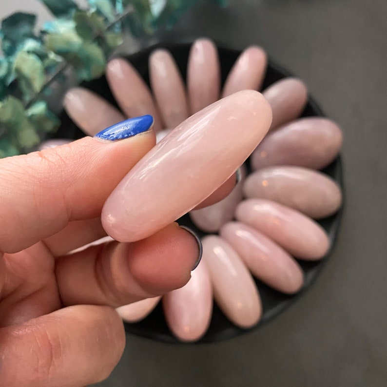 Pu&ograve; includere: Una collezione di pietre di quarzo rosa ovali e lisce in diverse tonalit&agrave; di rosa. Le pietre sono disposte in un motivo circolare su una superficie nera.