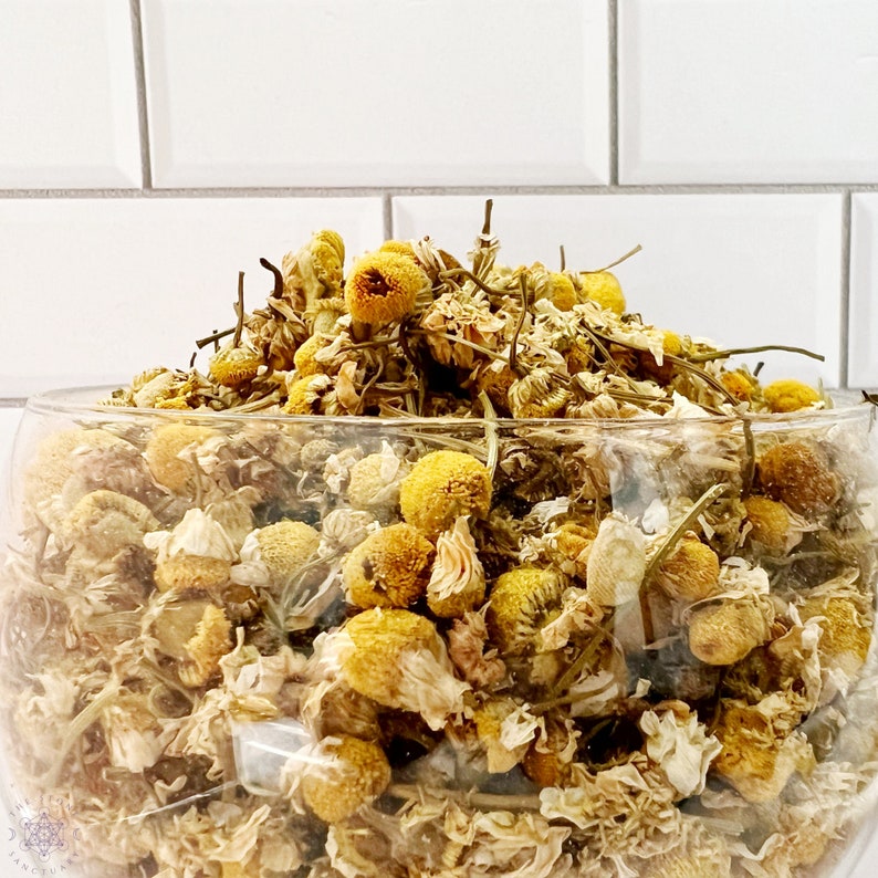 May include: A clear glass bowl filled with dried chamomile flowers. The flowers have yellow centers and white petals, with some stems visible. The bowl is set against a white tiled background, creating a clean and natural aesthetic.