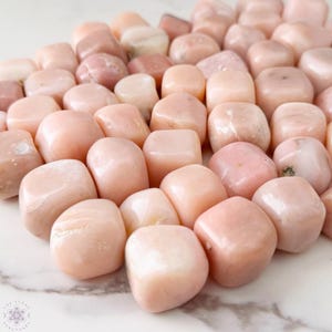 May include: A collection of smooth, tumbled pink opal stones. The stones vary in shades of pink, from pale blush to deeper rose, with a polished, glossy finish. The stones are arranged on a white and gray marble surface.