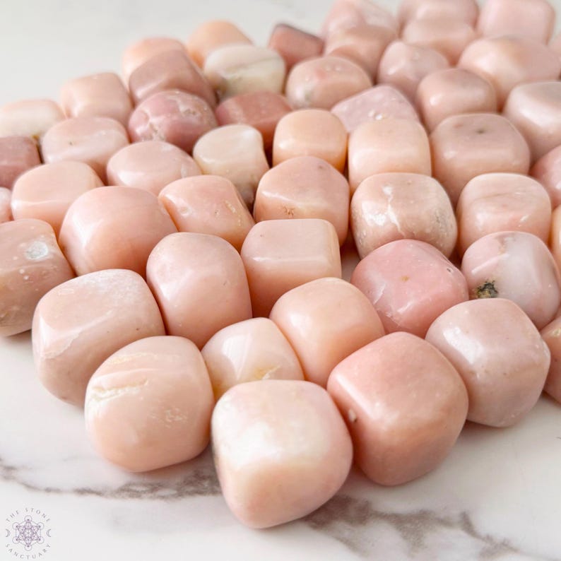 May include: A collection of smooth, tumbled pink opal stones. The stones vary in shades of pink, from pale blush to deeper rose, with a polished, glossy finish. The stones are arranged on a white and gray marble surface.