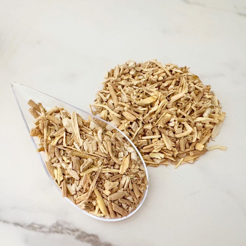 May include: A clear, teardrop-shaped scoop filled with light brown, chopped wood pieces, next to a pile of similar wood pieces. The wood pieces have a rough, splintered texture and vary in size and shape. The background is a white, marble-like surface.