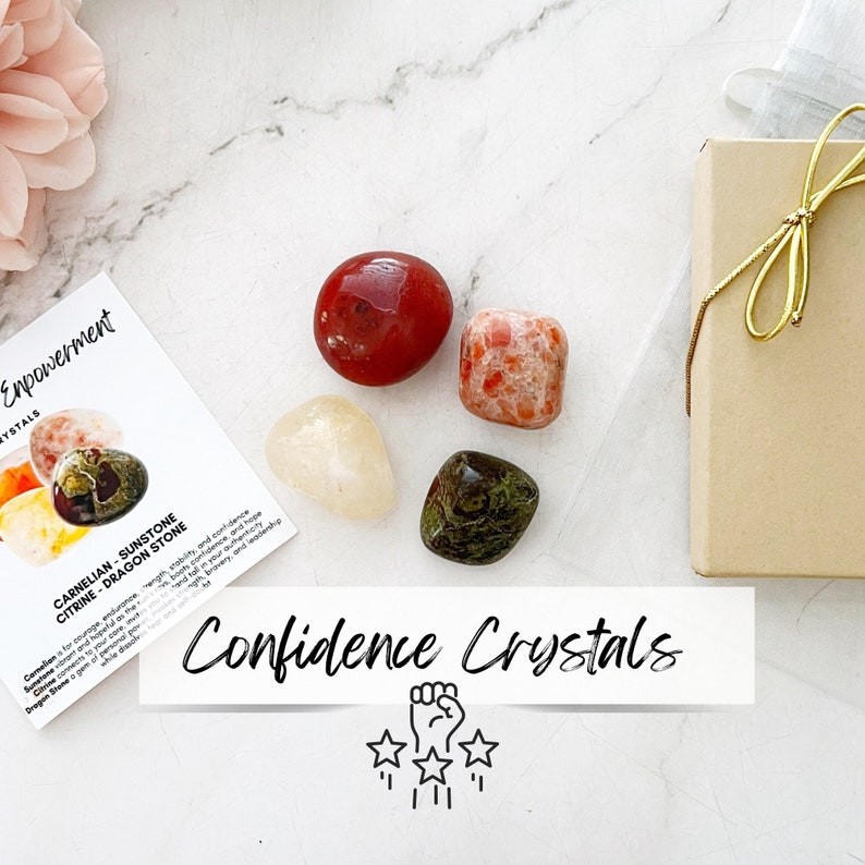 May include: A set of four crystals in a small box. The crystals are carnelian, sunstone, citrine, and dragon stone. The box is brown with a gold ribbon. The crystals are arranged in a circle with a white card in the center that says "Confidence Crystals".