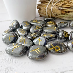 May include: A set of black hematite stones with gold runes inscribed on each one. The stones are tumbled and smooth.