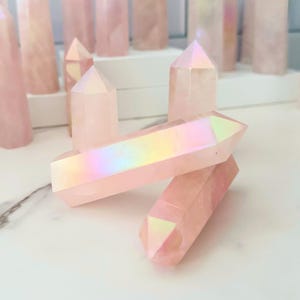 May include: A cluster of pink iridescent crystal points. The crystals are arranged in a group, with some overlapping. The crystals have a smooth, polished surface and a pointed tip. The crystals are a light pink colour with an iridescent sheen.
