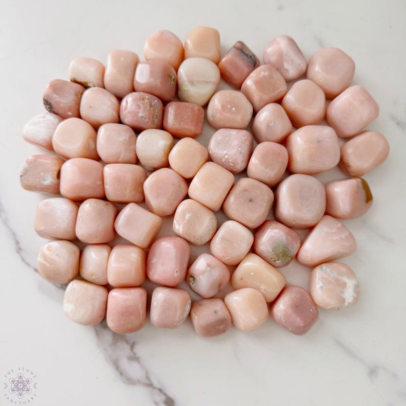 May include: A collection of tumbled pink opal stones. The stones vary in shades of pink, from pale blush to deeper rose, with smooth, rounded shapes. The stones are scattered on a white marble surface. The Stone Sanctuary logo is in the bottom left corner.