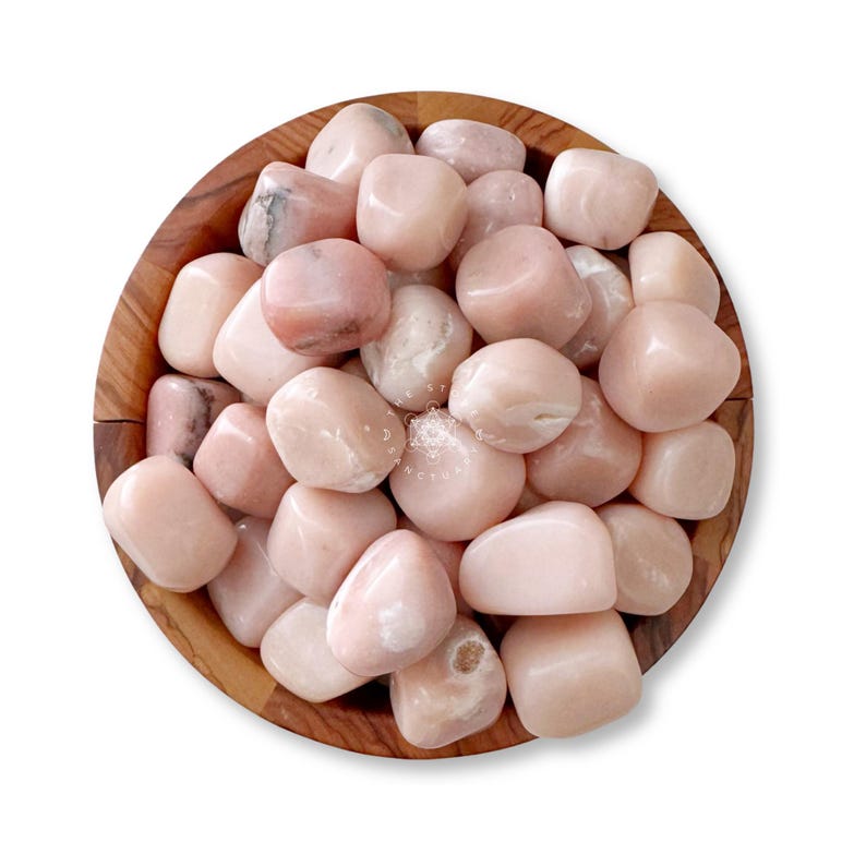 May include: A wooden bowl filled with smooth, tumbled pink stones. The stones vary in shade from pale to medium pink, with some showing subtle white marbling. The bowl is made of light brown wood, and the stones are the primary focus.