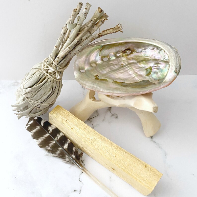 Puede incluir: Un manojo de salvia blanca, un palo santo y una concha de abul&oacute;n blanca sobre un soporte de madera. La concha es iridiscente y tiene una forma natural y org&aacute;nica.