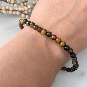 May include: A bracelet made of small, round, multi-colored beads. The beads are shades of brown, gold, red, and black. The bracelet is on a person's wrist.