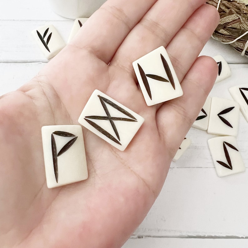 May include: Ivory-colored rune stones with dark brown etched symbols are displayed in a hand. The rectangular stones are scattered, with three prominent stones in focus. The runes are likely used for divination or spiritual practices.