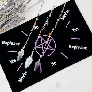 May include: Three crystal pendulums with silver chains hanging over a black velvet pendulum board with a pink pentagram and the words "Yes", "No", "Maybe", and "Rephrase" written in white.