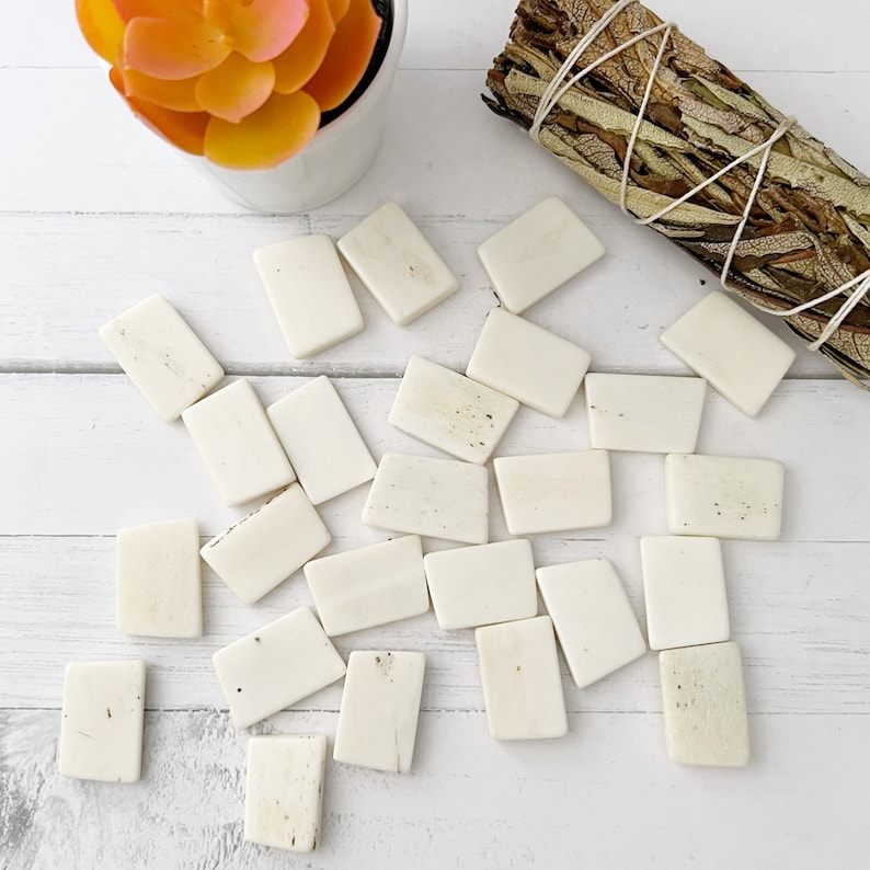 May include: A collection of off-white, rectangular bone tiles scattered on a white wooden surface. The tiles vary slightly in color and texture. A bundle of dried sage tied with twine and a small potted succulent are also visible.