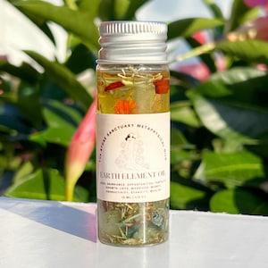 May include: A small glass bottle with a silver lid filled with a clear liquid and various herbs and crystals. The label reads "The Stone Sanctuary Metaphysical Oils Earth Element Oil" and lists benefits such as abundance, opportunity, growth, love, marriage, money, productivity, stability, and wealth. The bottle is 15 ml (1/2 oz).