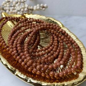 May include: A close-up of a pile of brown sandstone beads. The beads are round and have a speckled texture. They are arranged in a circular pattern on a gold-colored surface.