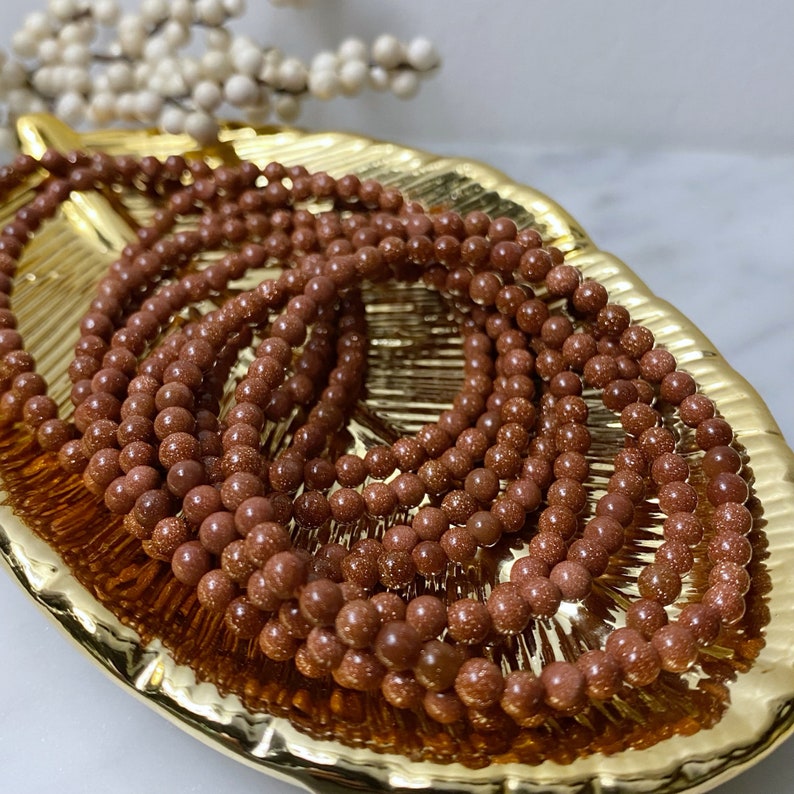 May include: A close-up of a pile of brown sandstone beads. The beads are round and have a speckled texture. They are arranged in a circular pattern on a gold-colored surface.