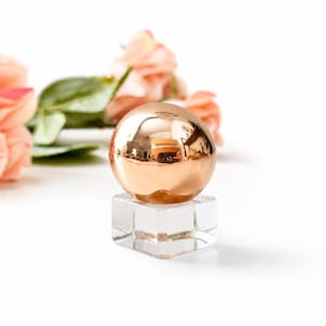 May include: A polished rose gold sphere resting on a clear, transparent glass cube base. The metallic surface of the sphere reflects the surrounding environment, while the base provides a stable, square foundation for the round object.