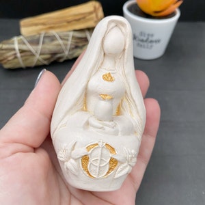 Puede incluir: Una figura de color crema de una mujer sentada con cabello largo y un velo. La figura sostiene un peque&ntilde;o recipiente y est&aacute; adornada con detalles dorados. La base presenta un emblema dorado y detalles florales. La figura se sostiene en una mano.