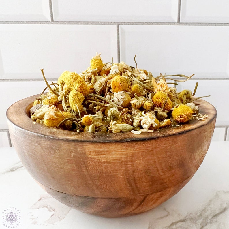 May include: A wooden bowl filled with dried chamomile flowers. The flowers are yellow and white, with green stems and leaves. The bowl is made of light brown wood and has a smooth finish. The Stone Sanctuary logo is visible in the bottom left corner.
