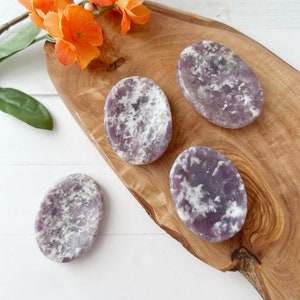 May include: Four purple and white Lepidolite palm stones on a wooden surface. The stones are smooth and oval shaped.