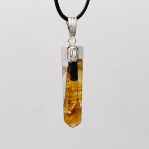 Puede incluir: Un colgante de cristal transparente con una tapa y una fianza de metal plateado, suspendido de un cordón negro. El cristal es de color ámbar claro con inclusiones marrones oscuras y una varilla de turmalina negra.