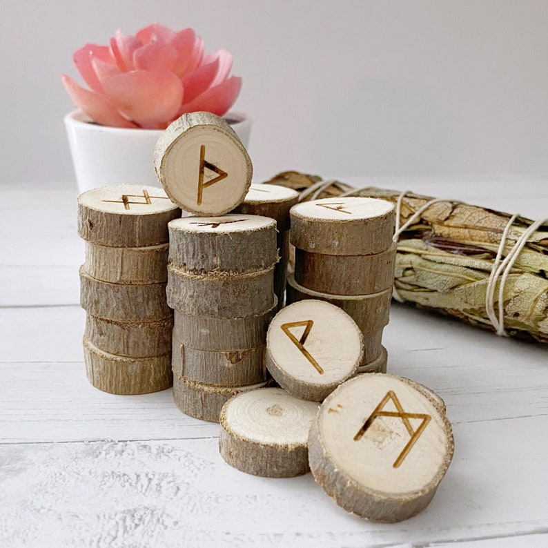 May include: A set of wooden rune tokens with different symbols carved into them. The tokens are stacked in a pile, with some of them arranged in a circle. The tokens are made of natural wood and have a rustic appearance.