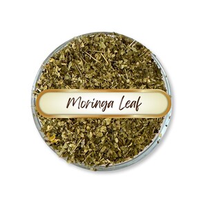 May include: A clear glass bowl filled with dried moringa leaves. A gold-colored label with the words "Moringa Leaf" in a handwritten style is centered in the image. The leaves are a mix of green and brown.
