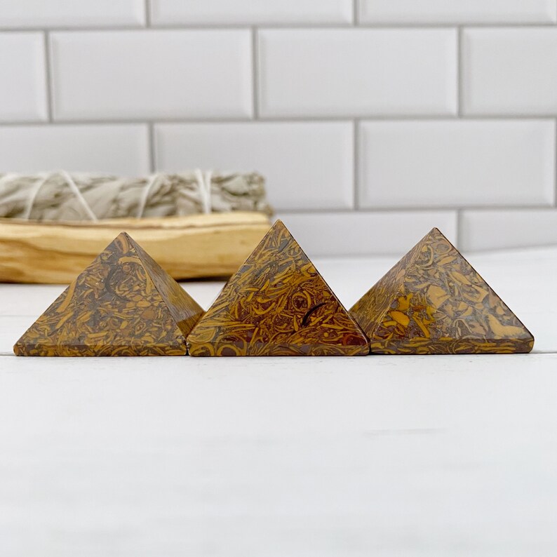 May include: Three brown and yellow patterned stone pyramids are arranged in a row. The pyramids have a textured surface and are set against a white background. A bundle of sage is in the background.