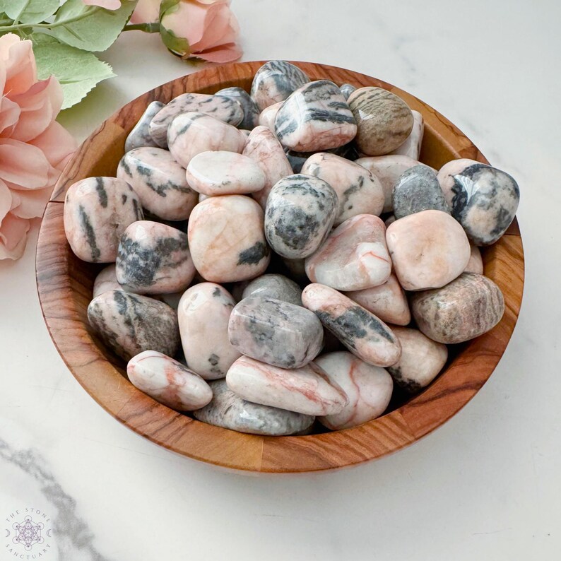May include: A wooden bowl filled with tumbled stones. The stones are a mix of pink, gray, and white, with various patterns and textures. The bowl is made of light brown wood. The stones are smooth and rounded, and appear to be polished.