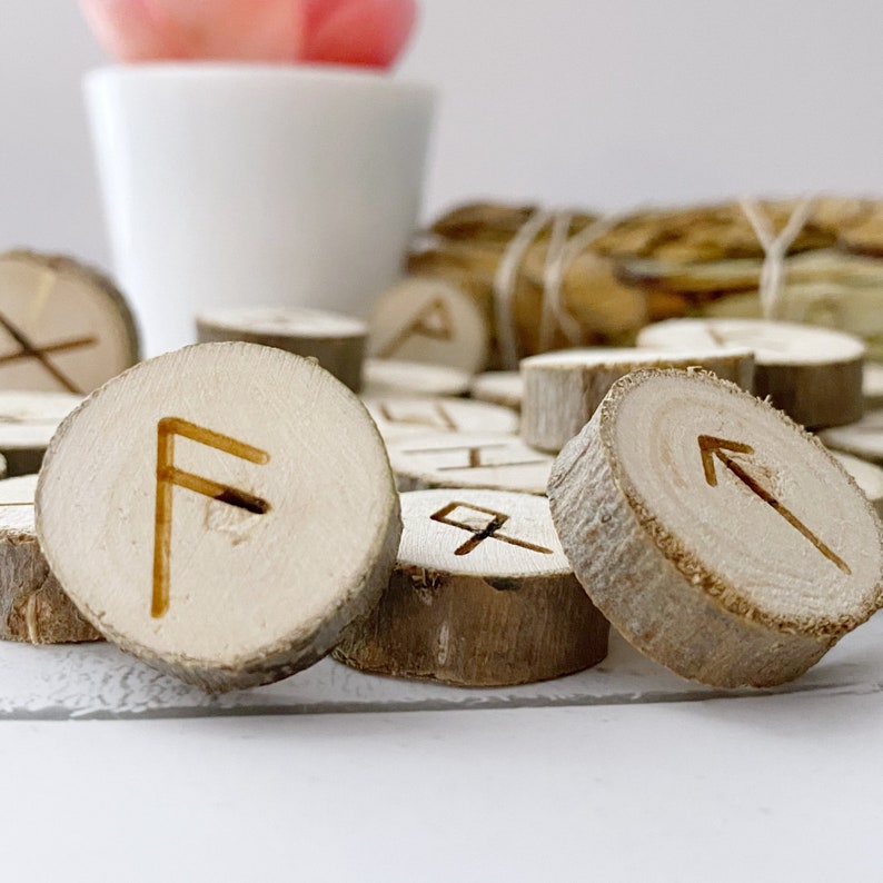 May include: Wooden runes with different symbols carved into them. The runes are arranged in a pile on a white surface.