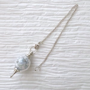 May include: A silver pendulum with a clear glass orb filled with white and blue crystals. The pendulum is attached to a silver chain.