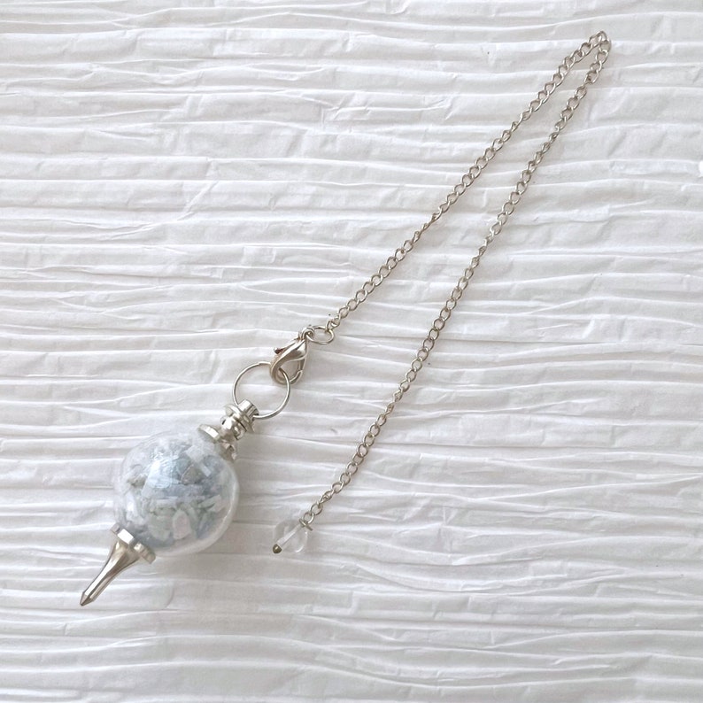 May include: A silver pendulum with a clear glass orb filled with white and blue crystals. The pendulum is attached to a silver chain.