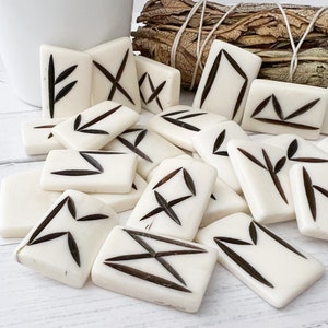 May include: A collection of off-white rune stones with dark brown markings. The rectangular and square stones are scattered, with some arranged in a pile. The runes are a mix of straight and angled lines, creating various symbols. A white ceramic container and a bundle of dried herbs are in the background.