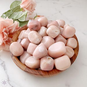 May include: A wooden bowl filled with smooth, polished pink opal stones. The stones vary in size and shape, with a soft, pearly sheen. A few pink roses and leaves are arranged near the bowl, adding a touch of natural beauty.