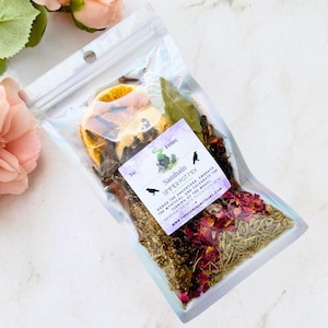 May include: A clear, iridescent bag filled with a simmer pot mix. The mix includes dried orange slices, bay leaves, cloves, and rose petals. A label reads "Samhain Simmer Pot Mix" with text about honoring ancestors and celebrating the turning of the wheel.